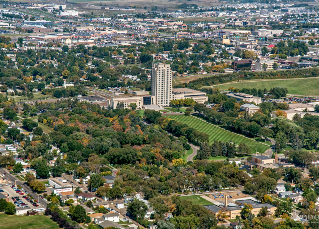 Aerial view of a city campus with a tall central tower surrounded by low-rise buildings and green lawns, amid a tree-filled urban area.